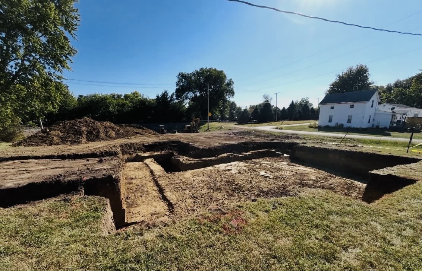 Basement excavation dug square with footing shelves cut clean, a farmhouse and road behind it, central Illinois