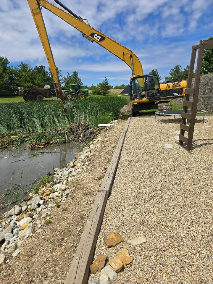 CAT 325D long-reach excavator pulling silt and cattails from a pond edge in North Peoria, Illinois, with a tractor and dump cart hauling soil behind